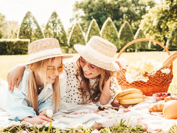 Moda elbisesi ve şapkası içinde iki güzel bayan. Dışarıda piknik yapan kaygısız kadınlar. Çimlerin üzerinde oturmuş meyve ve peynir yiyen pozitif modeller.