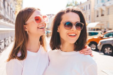 Son moda beyaz tişörtlü ve kot pantolonlu iki güzel, gülümseyen yeniyetme bayan sokak arka planında poz veren kaygısız kadınlar. Pozitif modeller eğleniyor, sarılıyor ve deliriyor.