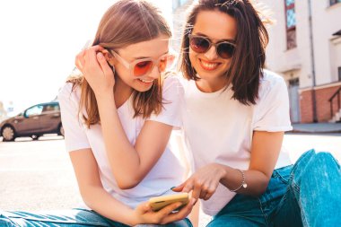 Son moda beyaz tişörtlü ve kot pantolonlu iki güzel gülümseyen yeniyetme kadın. Sokakta arka planda poz veren kaygısız kadınlar. Akıllı telefon uygulamaları kullanan, ekrana bakan pozitif modeller.