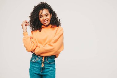 Afro kıvırcık saçlı, turuncu kapüşonlu ve kotlu güzel siyah kadın. Stüdyoda beyaz arka planda poz veren kaygısız seksi kadın. Bronzlaşmış ve neşeli