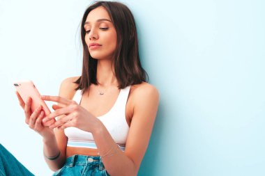 Beyaz formalı bluz ve kot pantolon giymiş güzel bir kadın. Seksi kaygısız neşeli model sabahının tadını çıkarıyor. Stüdyoda açık mavi duvarın yanında poz veren bir kadın var. Mobil uygulamalar kullanıyor.