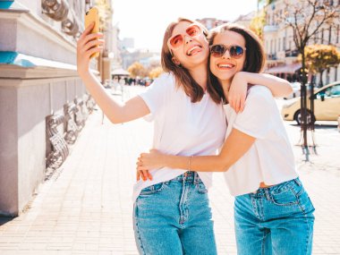 Son moda beyaz tişörtlü ve kot pantolonlu iki güzel gülümseyen yeniyetme kadın. Arka planda poz veren kaygısız kadınlar. Pozitif mankenler eğleniyor, sarılıyor.