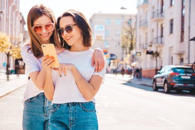 Son moda beyaz tişörtlü ve kot pantolonlu iki güzel gülümseyen yeniyetme kadın. Sokakta arka planda poz veren kaygısız kadınlar. Akıllı telefon uygulamaları kullanan, ekrana bakan pozitif modeller.