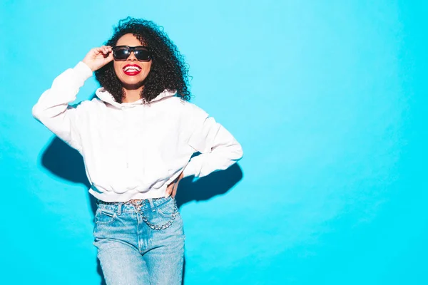 Beautiful black woman with afro curls hairstyle.Smiling model in trendy summer clothes. Sexy carefree female posing near blue wall in studio.In sunglasses with red lips
