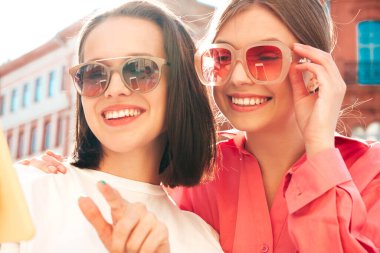 Son moda beyaz tişörtlü ve kot pantolonlu iki güzel gülümseyen yeniyetme kadın. Sokakta arka planda poz veren kaygısız kadınlar. Akıllı telefon uygulamaları kullanan, ekrana bakan pozitif modeller.