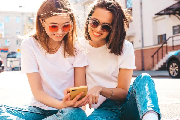 Son moda beyaz tişörtlü ve kot pantolonlu iki güzel gülümseyen yeniyetme kadın. Sokakta arka planda poz veren kaygısız kadınlar. Akıllı telefon uygulamaları kullanan, ekrana bakan pozitif modeller.