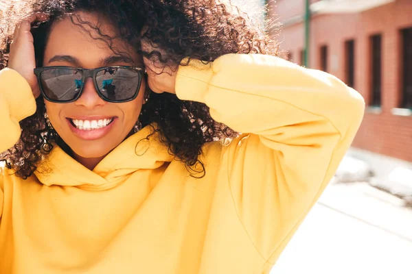 漂亮的黑人女人 有着非洲卷发的发型 穿着黄色帽衫的嬉皮士模特 穿着太阳镜的性感无忧无虑的女性在街上摆姿势 快乐而快乐 — 图库照片