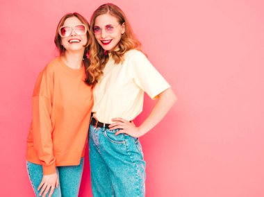 İki genç, güzel, gülümseyen yeniyetme kadın, modaya uygun yaz hippi kıyafetleri içinde. Stüdyoda pembe duvarın yanında poz veren kaygısız seksi kadınlar. Moda ve pozitif modeller eğleniyor. Neşeli ve mutlu