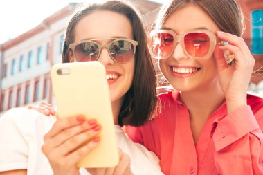 Son moda beyaz tişörtlü ve kot pantolonlu iki güzel gülümseyen yeniyetme kadın. Sokakta arka planda poz veren kaygısız kadınlar. Akıllı telefon uygulamaları kullanan, ekrana bakan pozitif modeller.