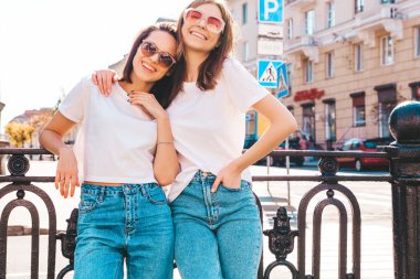 Son moda beyaz tişörtlü ve kot pantolonlu iki güzel, gülümseyen yeniyetme bayan sokak arka planında poz veren kaygısız kadınlar. Pozitif modeller eğleniyor, sarılıyor ve deliriyor.