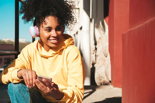 Beautiful black woman with afro curls hairstyle.Smiling model in yellow hoodie.Sexy carefree female enjoying listening music in wireless headphones.Posing on street background at sunset