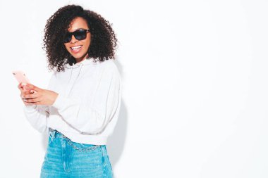 Saçları kıvırcık, güzel siyah bir kadın. Moda kıyafetleri içinde gülümseyen bir model. Stüdyoda izole edilmiş seksi, kaygısız kadın. Akıllı telefon uygulamaları kullanarak. Cep telefonu ekranına bakıyorum.