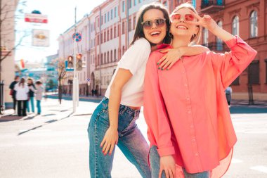 Son moda beyaz tişörtlü ve kot pantolonlu iki güzel, gülümseyen yeniyetme bayan sokak arka planında poz veren kaygısız kadınlar. Pozitif modeller eğleniyor, sarılıyor ve deliriyor.