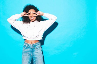 Saçları kıvırcık, güzel siyah bir kadın. Moda kıyafetleri içinde gülümseyen bir model. Stüdyoda mavi duvarın yanında poz veren kaygısız seksi kadın. Bronzlaşmış ve neşeli. Barış işareti nasıl?