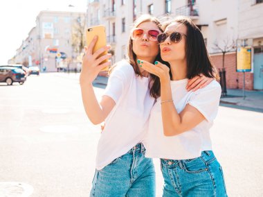 Son moda beyaz tişörtlü ve kot pantolonlu iki güzel gülümseyen yeniyetme kadın. Arka planda poz veren kaygısız kadınlar. Pozitif mankenler eğleniyor, sarılıyor.