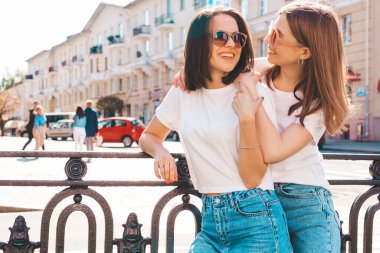 Son moda beyaz tişörtlü ve kot pantolonlu iki güzel, gülümseyen yeniyetme bayan sokak arka planında poz veren kaygısız kadınlar. Pozitif modeller eğleniyor, sarılıyor ve deliriyor.