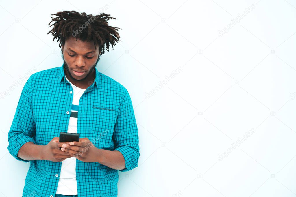 Handsome smiling hipster lambersexual model.Unshaven African man dressed in summer clothes.Fashion male with dreadlocks  posing near blue wall in studio.Looking at smartphone screen.Using apps 