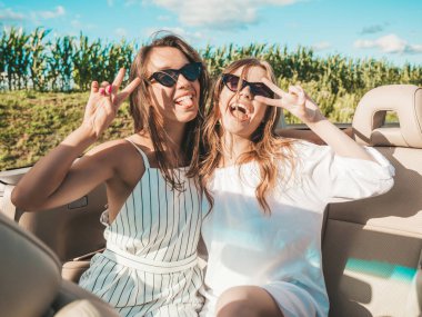 Üstü açık arabadaki iki güzel ve gülümseyen hippi kadının portresi. Cabriolet kullanan kaygısız seksi kadınlar. Pozitif modeller dışarıda güneş gözlüğü takıp eğleniyorlar. Keyifli yaz günleri