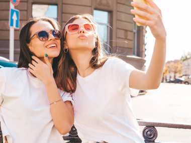 Son moda beyaz tişörtlü ve kot pantolonlu iki güzel gülümseyen yeniyetme kadın. Arka planda poz veren kaygısız kadınlar. Pozitif mankenler eğleniyor, sarılıyor.