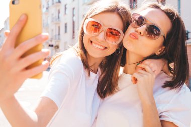 Son moda beyaz tişörtlü ve kot pantolonlu iki güzel gülümseyen yeniyetme kadın. Arka planda poz veren kaygısız kadınlar. Pozitif mankenler eğleniyor, sarılıyor.
