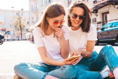 Son moda beyaz tişörtlü ve kot pantolonlu iki güzel gülümseyen yeniyetme kadın. Sokakta arka planda poz veren kaygısız kadınlar. Akıllı telefon uygulamaları kullanan, ekrana bakan pozitif modeller.