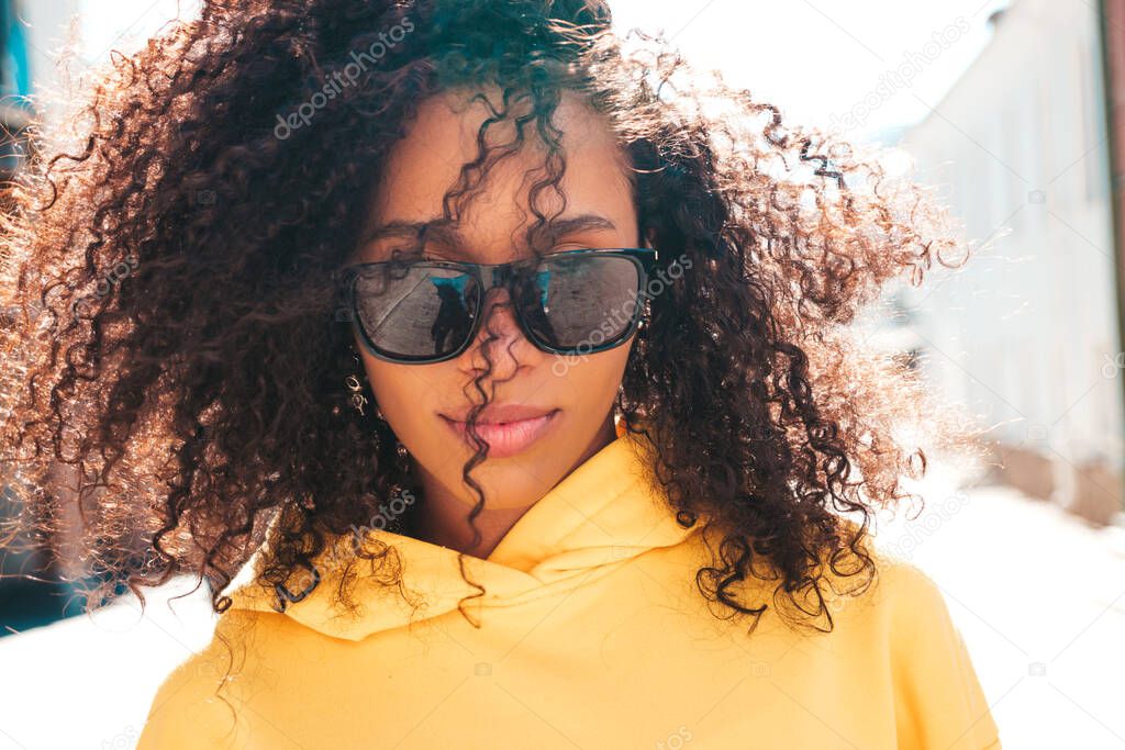 Beautiful black woman with afro curls hairstyle.Smiling hipster model in yellow hoodie. Sexy carefree female posing on the street background in sunglasses. Cheerful and happy