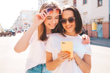 Son moda beyaz tişörtlü ve kot pantolonlu iki güzel gülümseyen yeniyetme kadın. Sokakta arka planda poz veren kaygısız kadınlar. Akıllı telefon uygulamaları kullanan, ekrana bakan pozitif modeller.