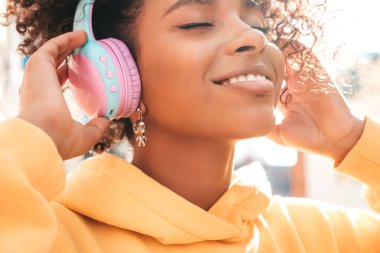 Afro kıvırcık saçlı güzel siyah kadın. Sarı kapüşonlu gülen manken. Kablosuz kulaklıkla müzik dinlemekten hoşlanan kaygısız kadın.