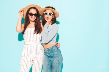 Son moda beyaz tişörtlü ve kot pantolonlu iki güzel gülümseyen hippi kadın stüdyoda açık mavi duvarın yanında poz veren kaygısız kadınlar. Şapkalı pozitif ve neşeli mankenler.