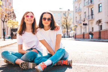 Son moda beyaz tişörtlü ve kot pantolonlu iki güzel, gülümseyen yeniyetme bayan sokak arka planında poz veren kaygısız kadınlar. Pozitif modeller eğleniyor, sarılıyor ve deliriyor.