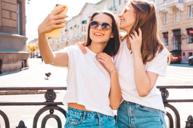 Son moda beyaz tişörtlü ve kot pantolonlu iki güzel gülümseyen yeniyetme kadın. Arka planda poz veren kaygısız kadınlar. Pozitif mankenler eğleniyor, sarılıyor.