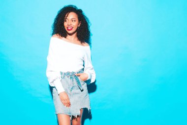 Saçları kıvırcık, güzel siyah bir kadın. Moda kıyafetleri içinde gülümseyen bir model. Stüdyoda mavi duvarın yanında poz veren kaygısız seksi kadın. Bronzlaşmış ve kıpkırmızı dudaklarla neşeli