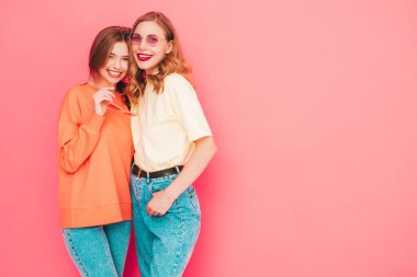 İki genç, güzel, gülümseyen yeniyetme kadın, modaya uygun yaz hippi kıyafetleri içinde. Stüdyoda pembe duvarın yanında poz veren kaygısız seksi kadınlar. Moda ve pozitif modeller eğleniyor. Neşeli ve mutlu