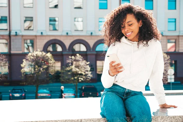Beautiful black woman with afro curls hairstyle.Smiling model in white hoodie. Sexy carefree female posing on the street background in sunglasses. Looking at smartphone screen, using apps