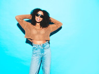 Saçları kıvırcık, güzel siyah bir kadın. Moda kıyafetleri içinde gülümseyen bir model. Stüdyoda mavi duvarın yanında poz veren kaygısız seksi kadın. Güneş gözlükleriyle bronzlaşmış ve neşeli