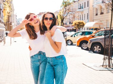 Son moda beyaz tişörtlü ve kot pantolonlu iki güzel, gülümseyen yeniyetme bayan sokak arka planında poz veren kaygısız kadınlar. Pozitif modeller eğleniyor, sarılıyor ve deliriyor.