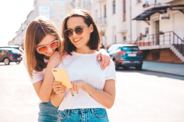 Son moda beyaz tişörtlü ve kot pantolonlu iki güzel gülümseyen yeniyetme kadın. Sokakta arka planda poz veren kaygısız kadınlar. Akıllı telefon uygulamaları kullanan, ekrana bakan pozitif modeller.