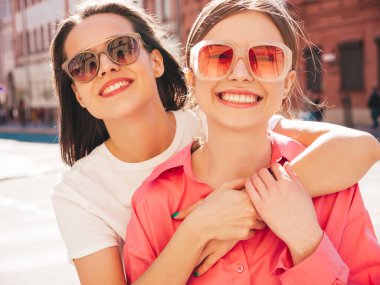 Son moda beyaz tişörtlü ve kot pantolonlu iki güzel, gülümseyen yeniyetme bayan sokak arka planında poz veren kaygısız kadınlar. Pozitif modeller eğleniyor, sarılıyor ve deliriyor.