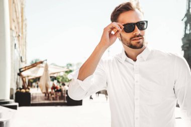 Yakışıklı, gülümseyen stil sahibi Lambersex modelinin portresi. Beyaz gömlek giymiş modern bir adam. Sokakta güneş gözlükleriyle poz veren modacı erkek. Gün batımında dışarıda. 