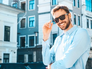 Yakışıklı, güler yüzlü, stil sahibi, lamberseksüel bir modelin portresi. Mavi gömlekli modern bir adam. Moda erkeği güneş gözlükleriyle gökdelenin yanında poz veriyor. Gün batımında dışarıda. 