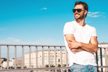 Yakışıklı, gülümseyen hipster Lambersex modelinin portresi. Beyaz tişört giymiş şık bir adam. Moda erkeği güneş gözlükleriyle mavi gökyüzünün arkasında poz veriyor. Kollar çapraz.