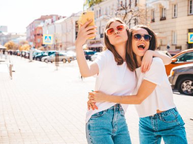 Son moda beyaz tişörtlü ve kot pantolonlu iki güzel gülümseyen yeniyetme kadın. Arka planda poz veren kaygısız kadınlar. Pozitif mankenler eğleniyor, sarılıyor.