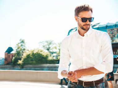 Yakışıklı, gülümseyen stil sahibi Lambersex modelinin portresi. Beyaz gömlek giymiş modern bir adam. Sokakta güneş gözlükleriyle poz veren modacı erkek. Gün batımında dışarıda. 