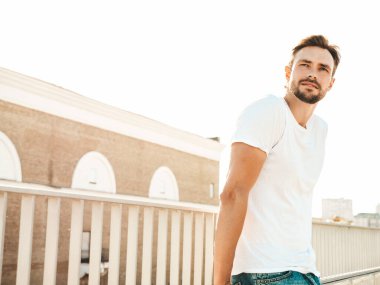 Yakışıklı, kendine güvenen hipster Lambersex modelinin portresi. Beyaz tişört ve kot giyen şık bir adam. Moda erkek, güneş gözlüklü, plajda çitlerin yanında poz veriyor.