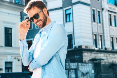 Yakışıklı, güler yüzlü, stil sahibi, lamberseksüel bir modelin portresi. Mavi gömlekli modern bir adam. Moda erkeği güneş gözlükleriyle gökdelenin yanında poz veriyor. Gün batımında dışarıda. 