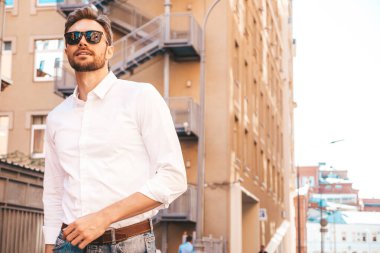 Yakışıklı, gülümseyen stil sahibi Lambersex modelinin portresi. Beyaz gömlek giymiş modern bir adam. Sokakta güneş gözlükleriyle poz veren modacı erkek. Gün batımında dışarıda. 