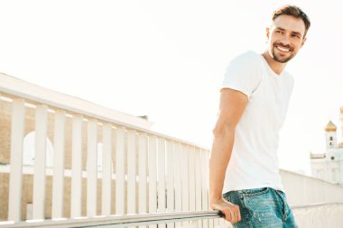 Yakışıklı, gülümseyen hipster Lambersex modelinin portresi. Beyaz tişört ve kot giyen şık bir adam. Moda erkek, güneş gözlüklü, plajda çitlerin yanında poz veriyor.