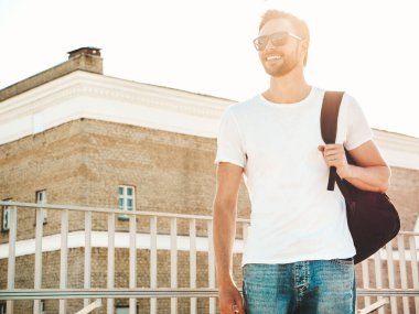 Yakışıklı, güler yüzlü, Lambersex mankeni. Beyaz tişört ve kot giyen şık bir adam. Moda erkek plajda çitlerin yanında güneş gözlüklü okul çantasıyla poz veriyor.