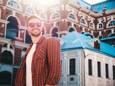 Yakışıklı, şık, havalı, havalı, lamberseksüel model. Kırmızı kareli gömlek giymiş modern bir adam. Moda erkeği güneş gözlükleriyle gökdelenin yanında poz veriyor. Gün batımında dışarıda. 