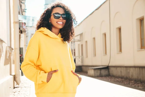 Beautiful black woman with afro curls hairstyle.Smiling hipster model in yellow hoodie. Sexy carefree female posing on the street background in sunglasses. Cheerful and happy